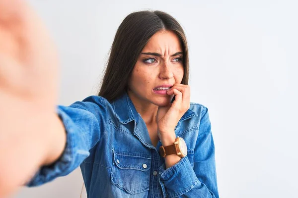 Beautiful woman wearing denim shirt make selfie by camera over isolated white background looking stressed and nervous with hands on mouth biting nails. Anxiety problem.