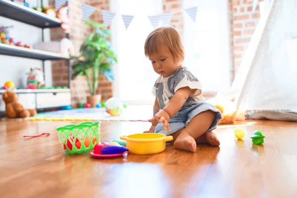 幼児は床に座ってプラスチック製のカトラリーや食べ物を使って食事をしています幼稚園のおもちゃの多くの周りに — ストック写真