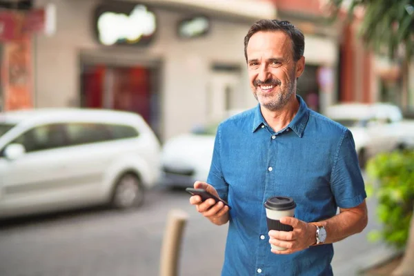 Middle age handsome businessman standing on the street using smartphone drinking take away coffee smiling