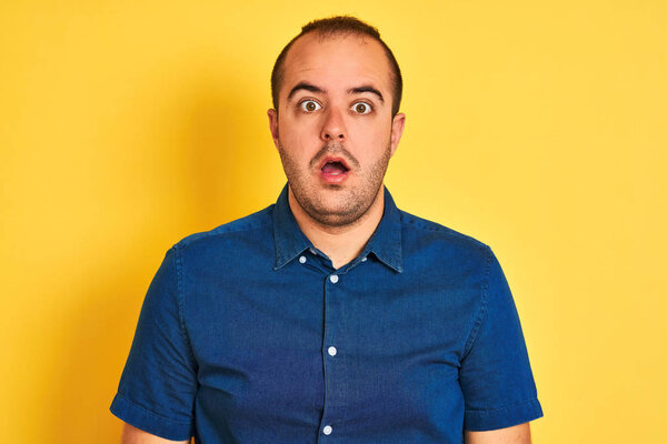 Young man wearing denim casual shirt standing over isolated yellow background afraid and shocked with surprise expression, fear and excited face.