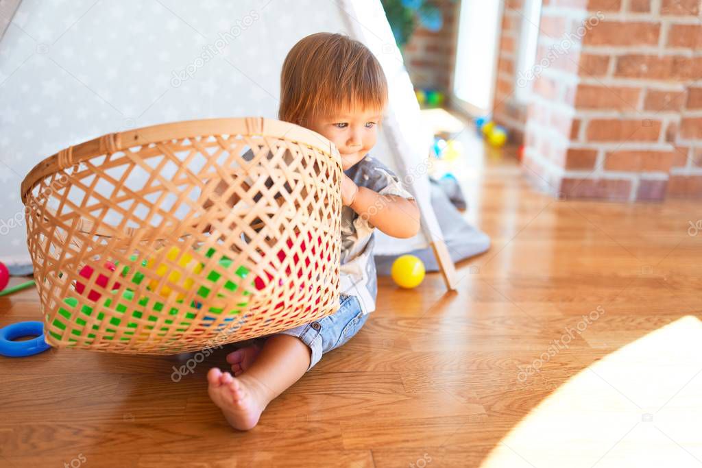 Adorable toddler sitting on the floor playing with wicker basket and balls around lots of toys at kindergarten