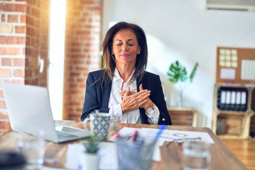 Middle age beautiful businesswoman working using laptop at the office smiling with hands on chest with closed eyes and grateful gesture on face. Health concept.