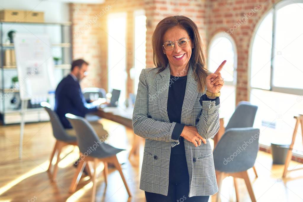 Middle age beautiful businesswoman wearing jacket and glasses standing at the office with a big smile on face, pointing with hand and finger to the side looking at the camera.