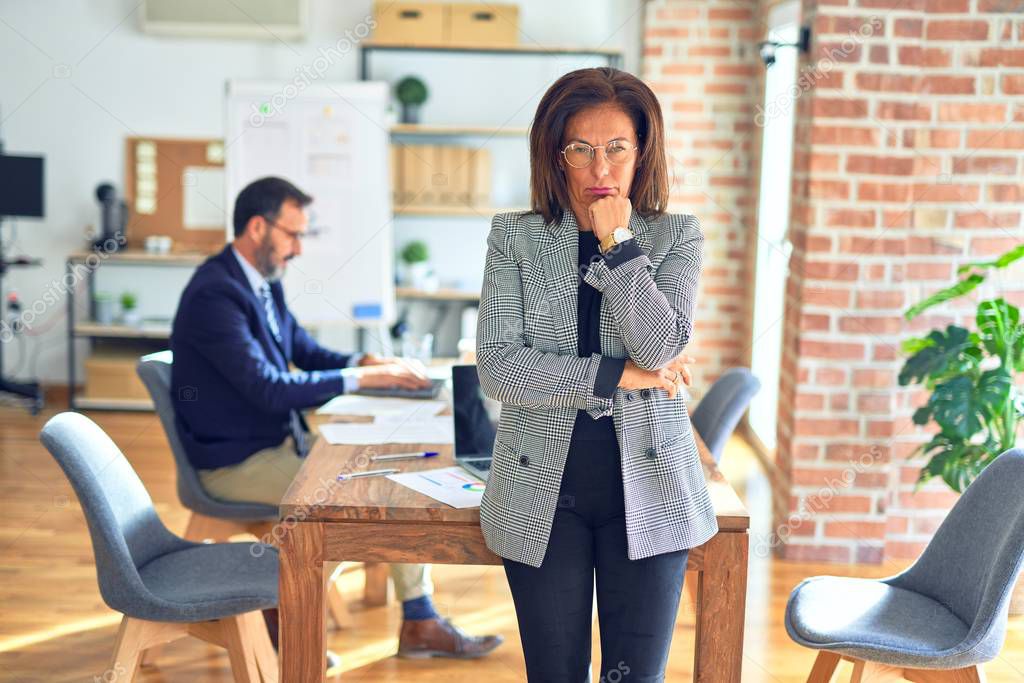 Middle age beautiful businesswoman wearing jacket and glasses standing at the office thinking looking tired and bored with depression problems with crossed arms.