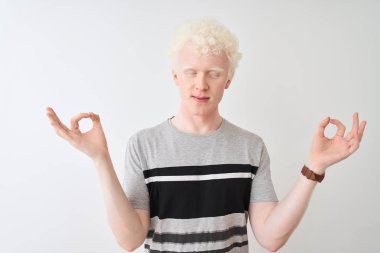 İzole edilmiş beyaz arka planda duran sıradan tişört giyen genç albino sarışın adam rahatla ve gözleri kapalı meditasyon hareketi yaparken parmaklarınla gülümse. Yoga konsepti.