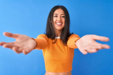 Young beautiful chinese woman wearing orange t-shirt standing over isolated blue background looking at the camera smiling with open arms for hug. Cheerful expression embracing happiness.