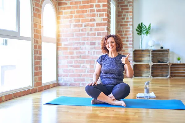 Middle age beautiful sportswoman wearing sportswear sitting on mat practicing yoga at home doing happy thumbs up gesture with hand. Approving expression looking at the camera with showing success.