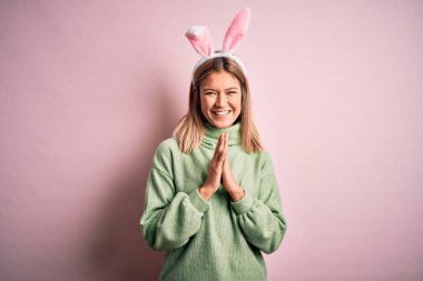 Paskalya tavşanı kulakları takan genç ve güzel bir kadın pembe arka planda durup elleriyle dua ederek af diliyor kendinden emin bir şekilde gülümsüyor..