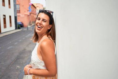 Güzel kız beyaz duvara yaslanıyor, genç arkadaş canlısı kadın mutlu bir yaz gününde gülümsüyor.