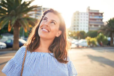 Genç ve güzel bir kadın mutlu bir şekilde gülümsüyor güneşli bir yaz gününde şehir sokaklarında yürüyor.