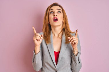 Young caucasian business woman wearing a suit over isolated pink background amazed and surprised looking up and pointing with fingers and raised arms.