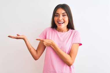 İzole edilmiş beyaz arka plan üzerinde duran pembe günlük tişört giyen genç ve güzel bir kadın kameraya hayret ediyor ve gülümsüyor..
