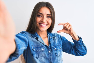 Kot gömlek giyen güzel bir kadın, izole edilmiş beyaz arka planda kamerayla selfie çekiyor gülümsüyor ve el hareketleriyle küçük boy işareti yapıyor, parmaklarıyla bakıyor ve kameraya bakıyor. Ölçü kavramı.
