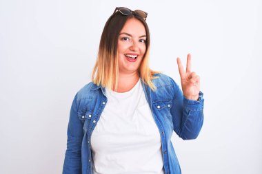 İzole edilmiş beyaz arka plan üzerinde duran kot gömlek giyen güzel bir kadın iki numaralı parmaklarıyla işaret ederken kendinden emin ve mutlu bir şekilde gülümsüyor..