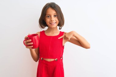 Kırmızı çay bardağını tutan güzel bir kız çocuğu. İzole edilmiş beyaz arka plan üzerinde duruyor. Şaşırmış yüz ifadesiyle kendini işaret ediyor.