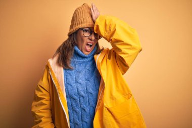 Middle age woman wearing yellow raincoat and winter hat over isolated background surprised with hand on head for mistake, remember error. Forgot, bad memory concept. clipart