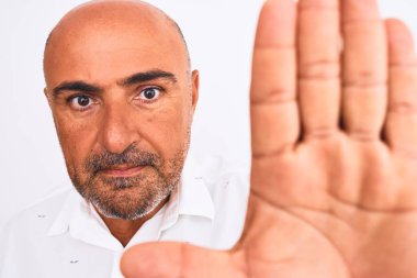 Orta yaşlı, şık gömlekli yakışıklı bir adam izole edilmiş beyaz arka planda duruyor. Açık elle dur işareti yapıyor. Ciddi ve kendinden emin bir ifadeyle, savunma jesti yapıyor.