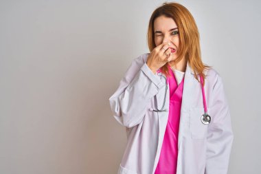 Redhead caucasian doctor woman wearing pink stethoscope over isolated background smelling something stinky and disgusting, intolerable smell, holding breath with fingers on nose. Bad smells concept.