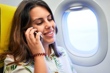 Havaalanında oturan genç yolcu kadın. Pencereden gökyüzü manzaralı. Akıllı telefon kullanarak arıyor.