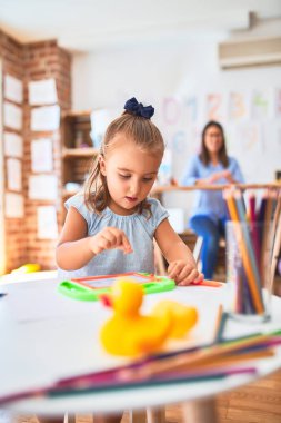 Beyaz kız çocuk, bayan öğretmenle oyun oynayıp öğreniyor. Anne ve kızı oyun odasında oyuncakların etrafında manyetik karatahtaya resim çiziyorlar.