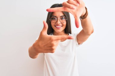 İzole edilmiş beyaz arka planda günlük tişört ve gözlük takan genç ve güzel bir kadın gülümsüyor ve mutlu yüzlü elleri ve parmakları olan bir çerçeve yapıyor. Yaratıcılık ve fotoğrafçılık kavramı.