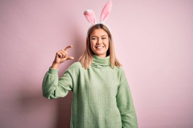 Paskalya tavşanı kulakları takan genç ve güzel bir kadın pembe arka planda duruyor gülümsüyor ve kendinden emin bir şekilde el hareketi yapıyor parmaklarıyla işaret yapıyor ve kameraya bakıyor. Ölçü kavramı.