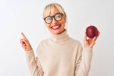 Elma tutan orta yaşlı bir kadın soyutlanmış beyaz arka planda duruyor. Elleri ve parmakları kenarda mutlu bir şekilde işaret ediyor.