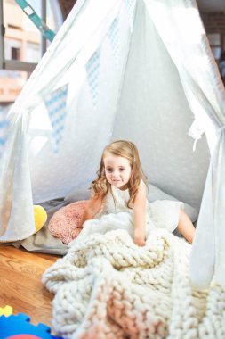 Anaokulunun içinde battaniyenin üzerinde oturan sevimli sarışın çocuk.