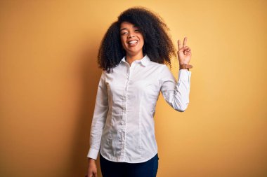 Afro saçlı, genç ve güzel Afro-Amerikan zarif bir kadın sarı arka planda duruyor ve parmaklarını iki numarayı gösterirken kendinden emin ve mutlu bir şekilde gülümsüyor..