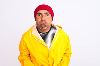 Yağmurluk giyen ve yünlü şapka takan orta yaşlı bir adam izole edilmiş beyaz arka planda durup komik yüzlü yanakları şişiriyor. Havayla şişmiş ağız, çılgın bir ifade..