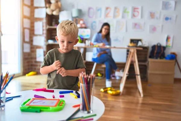 若い白人の子供は教師と一緒に学校で遊んでいる 母と息子は遊び場で色鉛筆で絵を描く若い女性は机の上に座っている — ストック写真