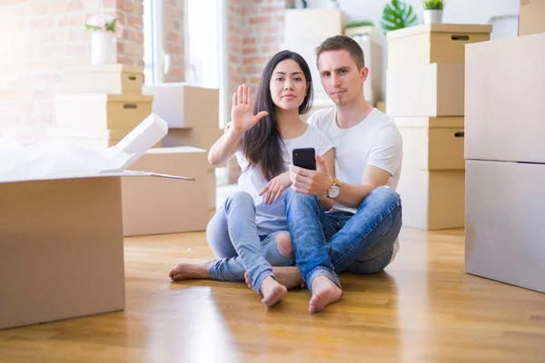 Beautiful couple sitting on the floor using smartphone at new home around cardboard boxes with open hand doing stop sign with serious and confident expression, defense gesture