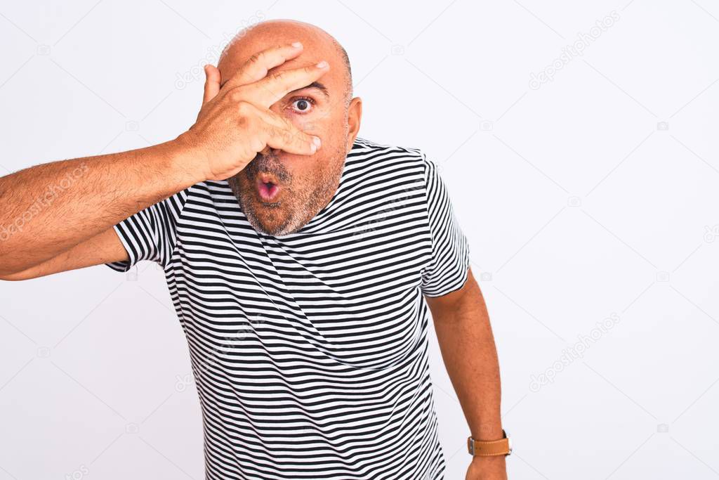 Middle age handsome man wearing striped navy t-shirt over isolated white background peeking in shock covering face and eyes with hand, looking through fingers with embarrassed expression.