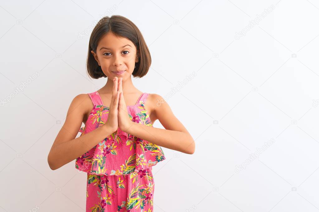 Young beautiful child girl wearing pink floral dress standing over isolated white background praying with hands together asking for forgiveness smiling confident.