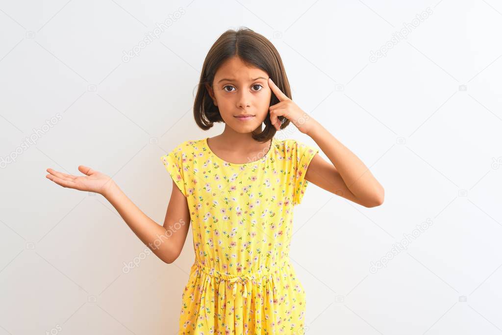 Young beautiful child girl wearing yellow floral dress standing over isolated white background confused and annoyed with open palm showing copy space and pointing finger to forehead. Think about it.
