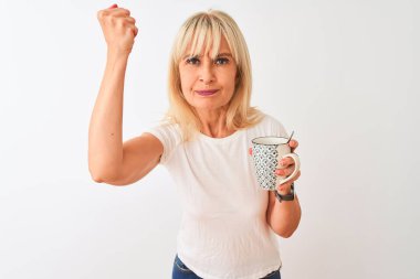 Orta yaşlı bir kadın, izole edilmiş beyaz arka planda dikilmiş bir fincan kahve içiyor sinirlenmiş ve sinirlenmiş bir şekilde bağırıyor, çıldırmış ve el kaldırarak bağırıyor.