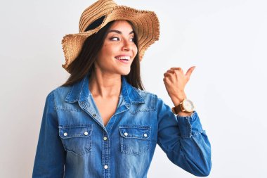 Kot gömlek ve şapka giyen genç güzel bir kadın izole edilmiş beyaz arka planda gülümsüyor ve başparmağıyla yan tarafa bakıyor..