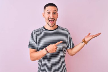 Çizgili tişört giyen genç yakışıklı adam pembe izole edilmiş arka planda durup hayret ediyor ve kameraya gülümsüyor..