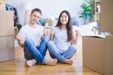 Young beautiful couple sitting on the floor at new home around cardboard boxes celebrating surprised and amazed for success with arms raised and open eyes. Winner concept.