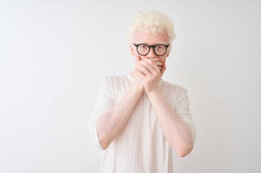 Tişört giyen ve gözlüklü genç albino bir adam izole edilmiş beyaz arka planda dikiliyor. Ağzını yanlışlıkla ellerle kapatıyor. Gizli konsept.