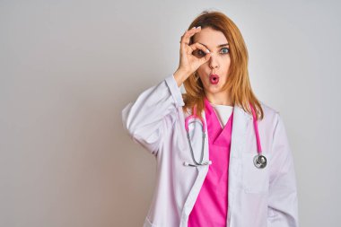 Redhead caucasian doctor woman wearing pink stethoscope over isolated background doing ok gesture shocked with surprised face, eye looking through fingers. Unbelieving expression.