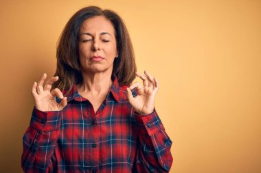 Orta yaşlı, güzel bir kadın, izole edilmiş sarı arka planda dikilmiş, rahatlamış ve gözleri kapalı meditasyon hareketi yaparken parmaklarıyla gülümsüyor. Yoga konsepti.