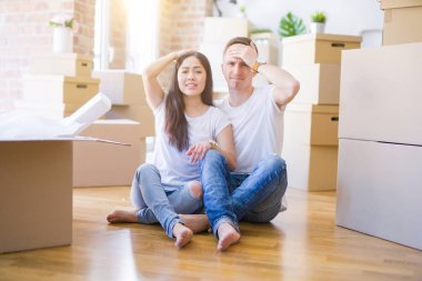 Young beautiful couple sitting on the floor at new home around cardboard boxes stressed with hand on head, shocked with shame and surprise face, angry and frustrated. Fear and upset for mistake.
