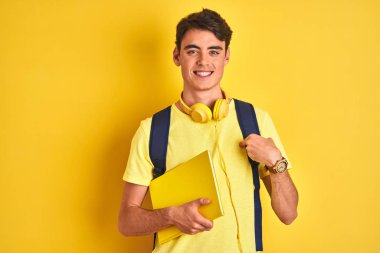Teenager boy wearing headphones and backpack reading a book over isolated background with surprise face pointing finger to himself