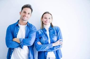 İzole edilmiş beyaz arka plan üzerinde duran kot gömlek giyen genç ve güzel bir çift kameraya bakarken kollarını kavuşturup gülümsüyor. Pozitif kişi.