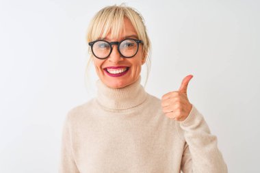 Balıkçı yaka kazak giyen ve gözlüklü orta yaşlı bir kadın izole edilmiş beyaz arka planda mutlu bir el hareketi yapıyor. Kameraya bakıp başarıyı gösteren ifade onaylanıyor.