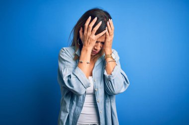 Orta yaşlı, güzel bir kadın, izole edilmiş mavi arka plan üzerinde dikilip acı ve migren yüzünden çaresiz ve stresli bir şekilde acı çekiyor. Ellerini başının üstüne koy..