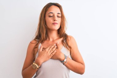 Genç kızıl saçlı kadın, günlük tişört giyiyor, beyaz tenli arka planda, gülümsüyor, elleri göğsünde, gözleri kapalı ve yüzünde minnet dolu bir jest var. Sağlık kavramı.