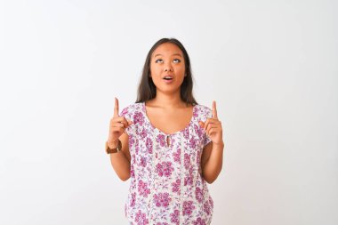 İzole edilmiş beyaz arka planda dikilen günlük çiçekli tişört giyen genç Çinli kadın şaşırmış ve şaşırmış bir şekilde yukarı bakıp parmaklarıyla işaret ediyor ve kollarını kaldırıyordu..