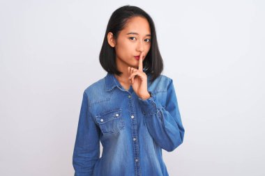 Young beautiful chinese woman wearing denim shirt standing over isolated white background asking to be quiet with finger on lips. Silence and secret concept.